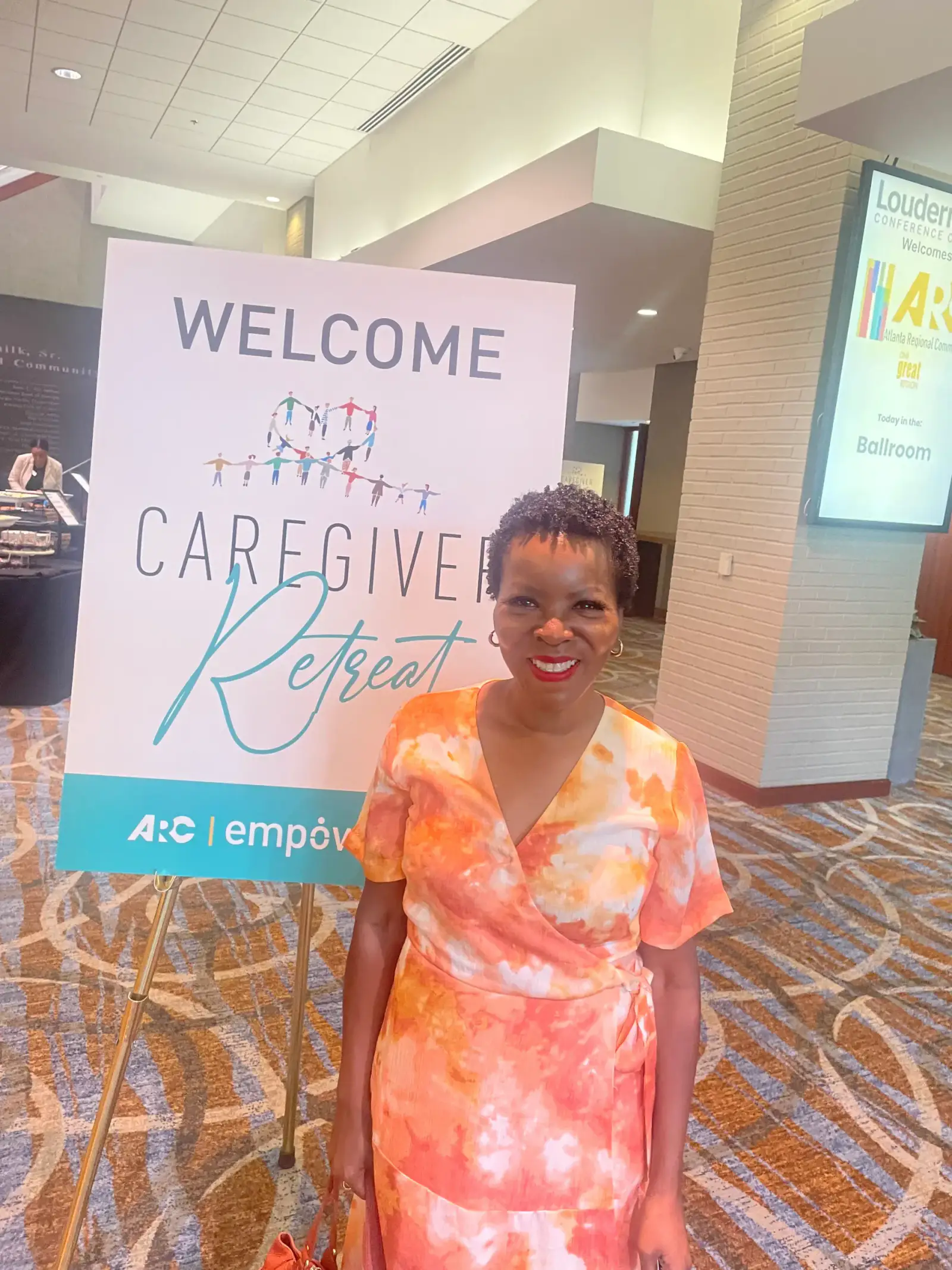 Shawn Caprice Houston smiling in front of a Caregiver Retreat welcome sign