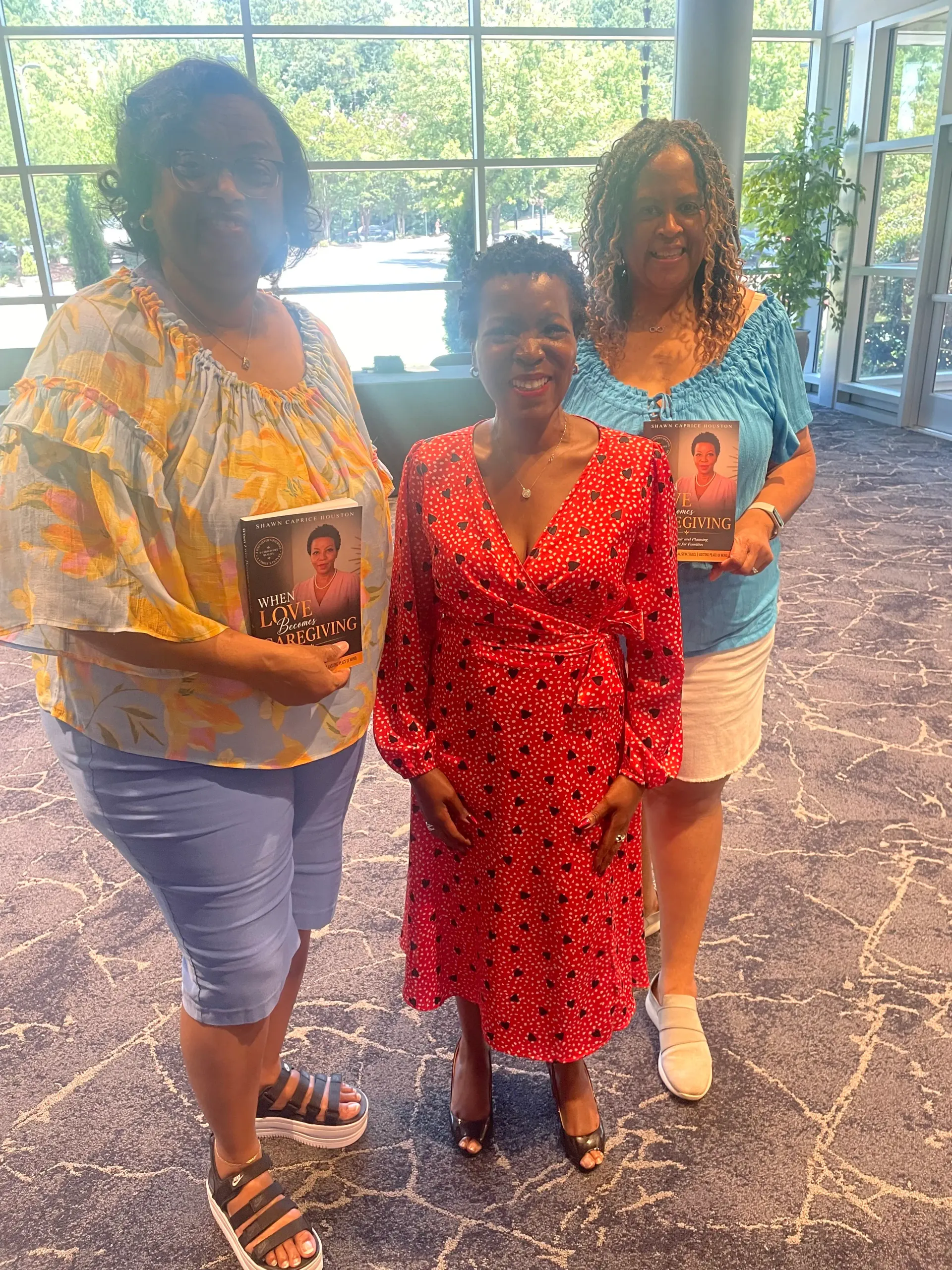 Shawn Caprice Houston smiling between two community members, each holding a copy of When Love Becomes Caregiving at an indoor gathering